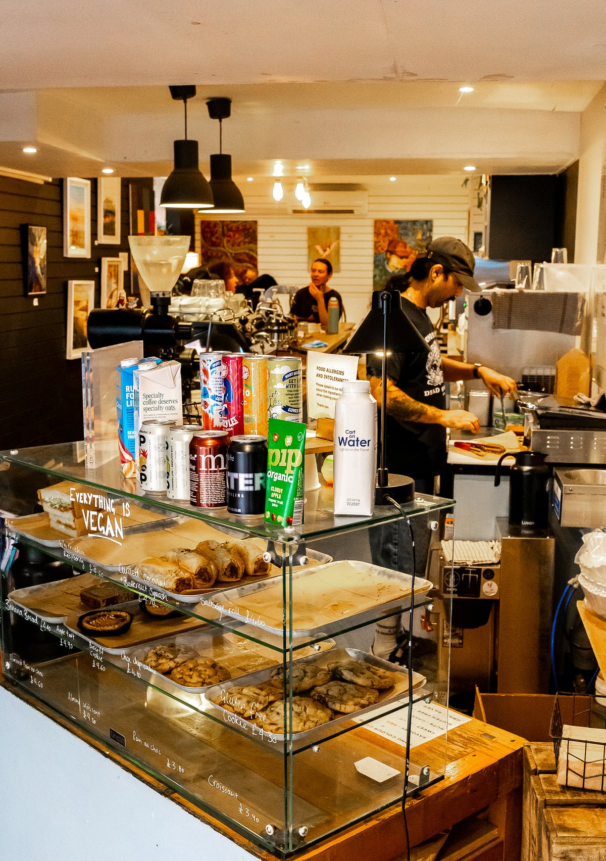 A busy cafe interior in Brighton with a glass display case holding vegan pastries on the counter, surrounded by drinks and a barista working behind the scene.
