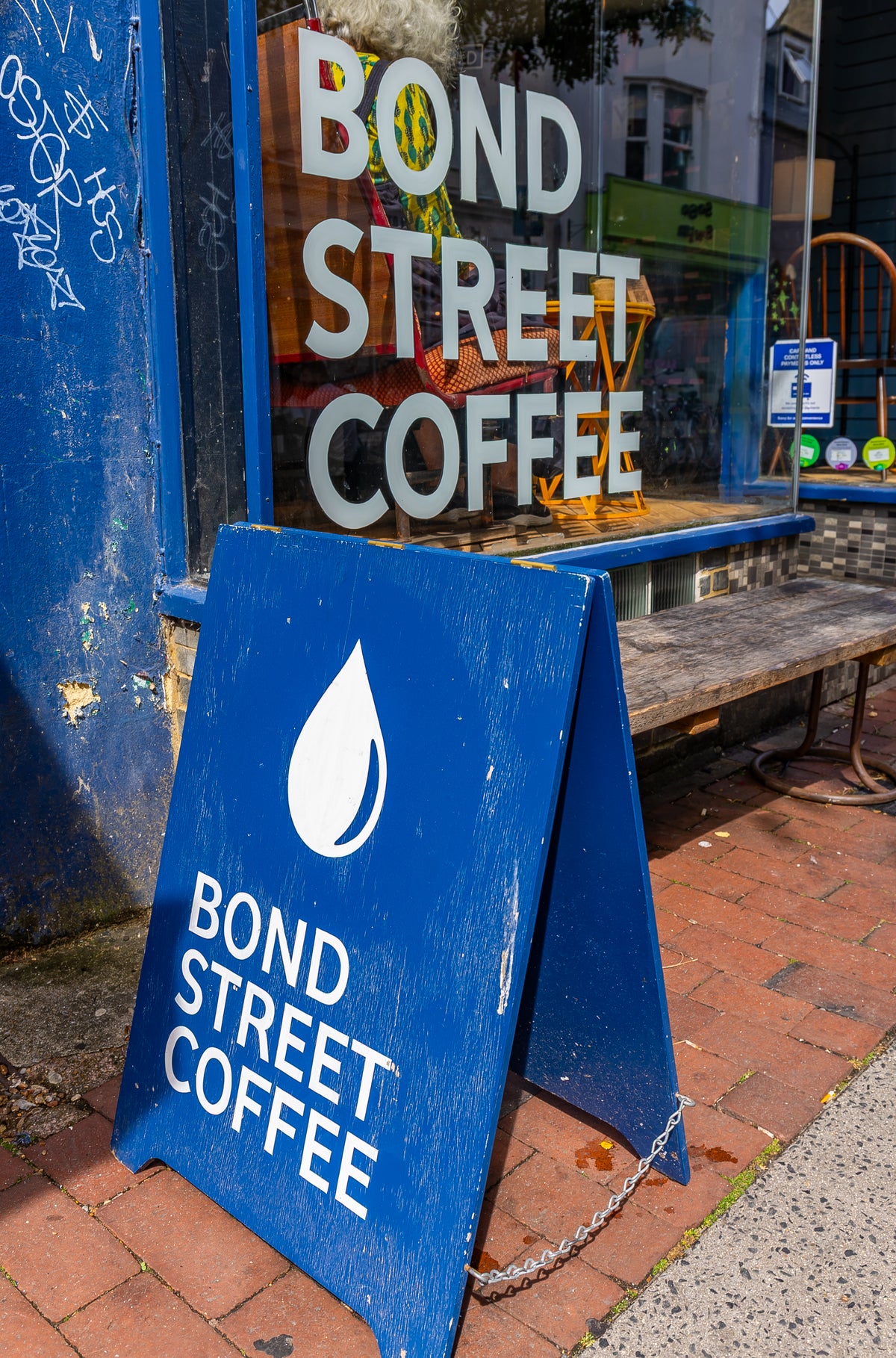 A blue A-frame sidewalk sign for Bond Street Coffee Company stands on a brick sidewalk in the Brighton North Laine with the coffee shop window, which also features large white 'BOND STREET COFFEE' lettering on a glass pane.