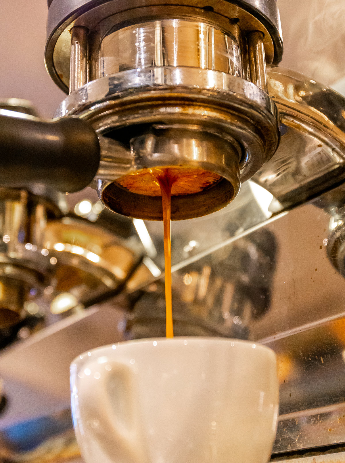 A close-up shot of single origin espresso being poured from a metal portafilter into a white ceramic cup, with steam rising in the background.