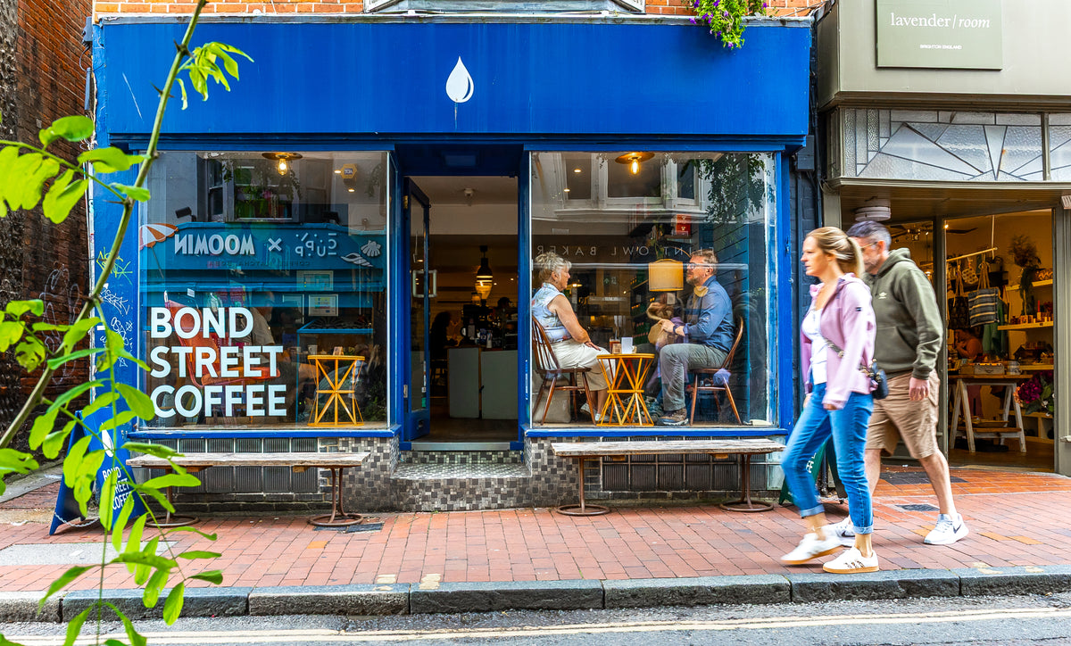 A blue storefront for Bond Street Coffee with large windows, seated people inside, and two pedestrians walking in front on a brick sidewalk.