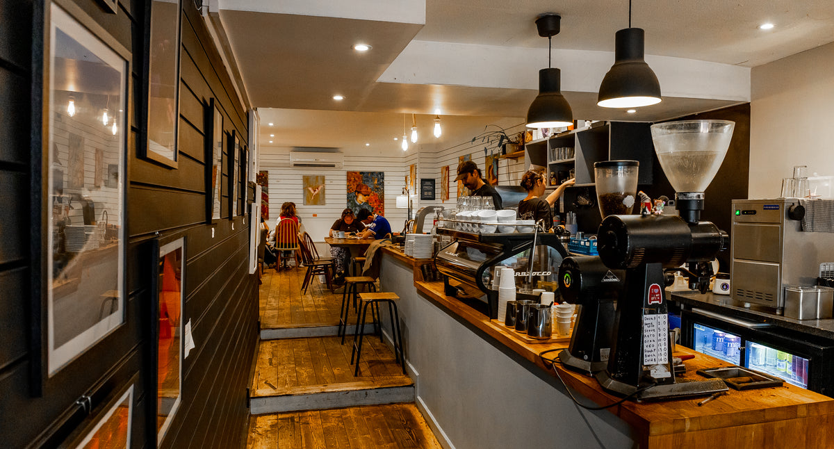 A cozy, rustic speciality coffee shop interior with a wooden counter, espresso machines, hanging black pendant lights, and patrons seated in the background.