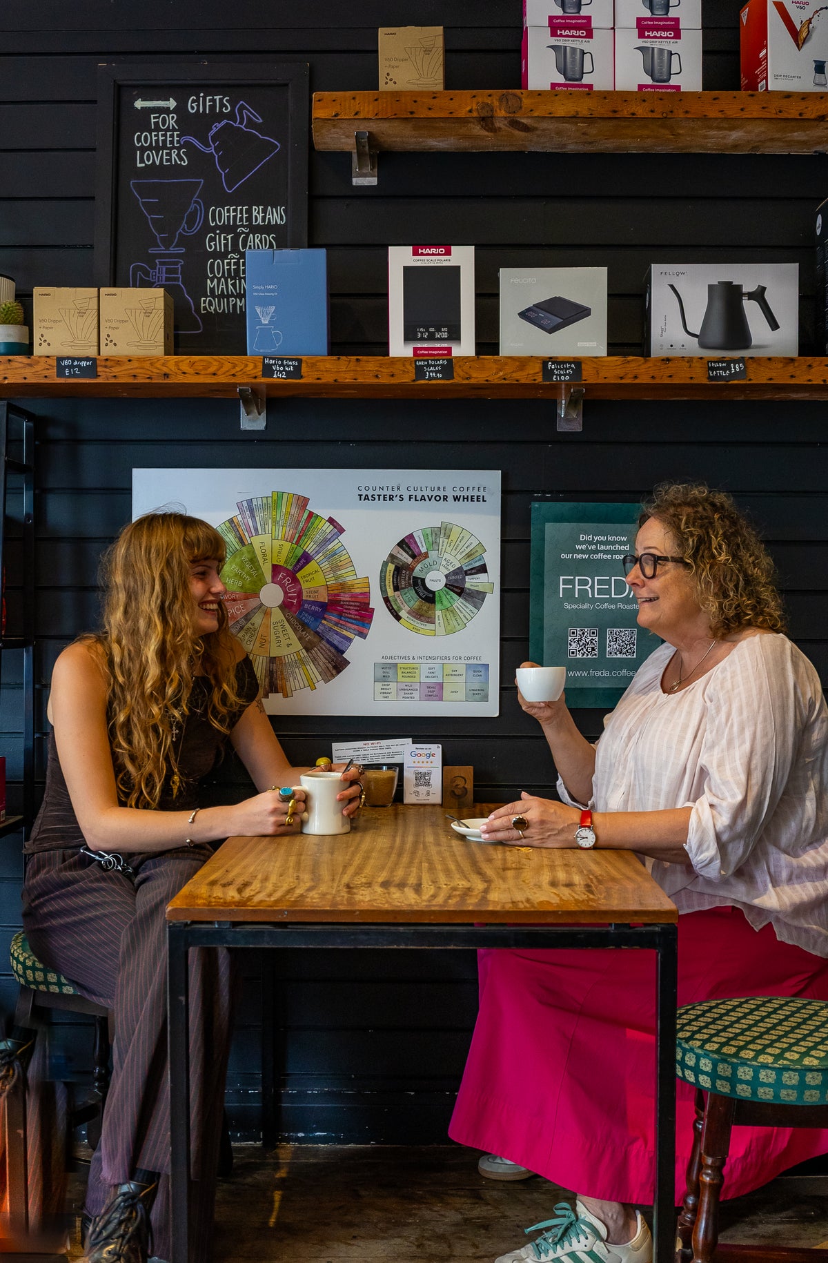 Two women sitting at a wooden table in a coffee shop, holding white mugs and engaged in conversation in Brighton. The background features dark wood shelves with speciality coffee-related products and a colorful player model poster on the wall.