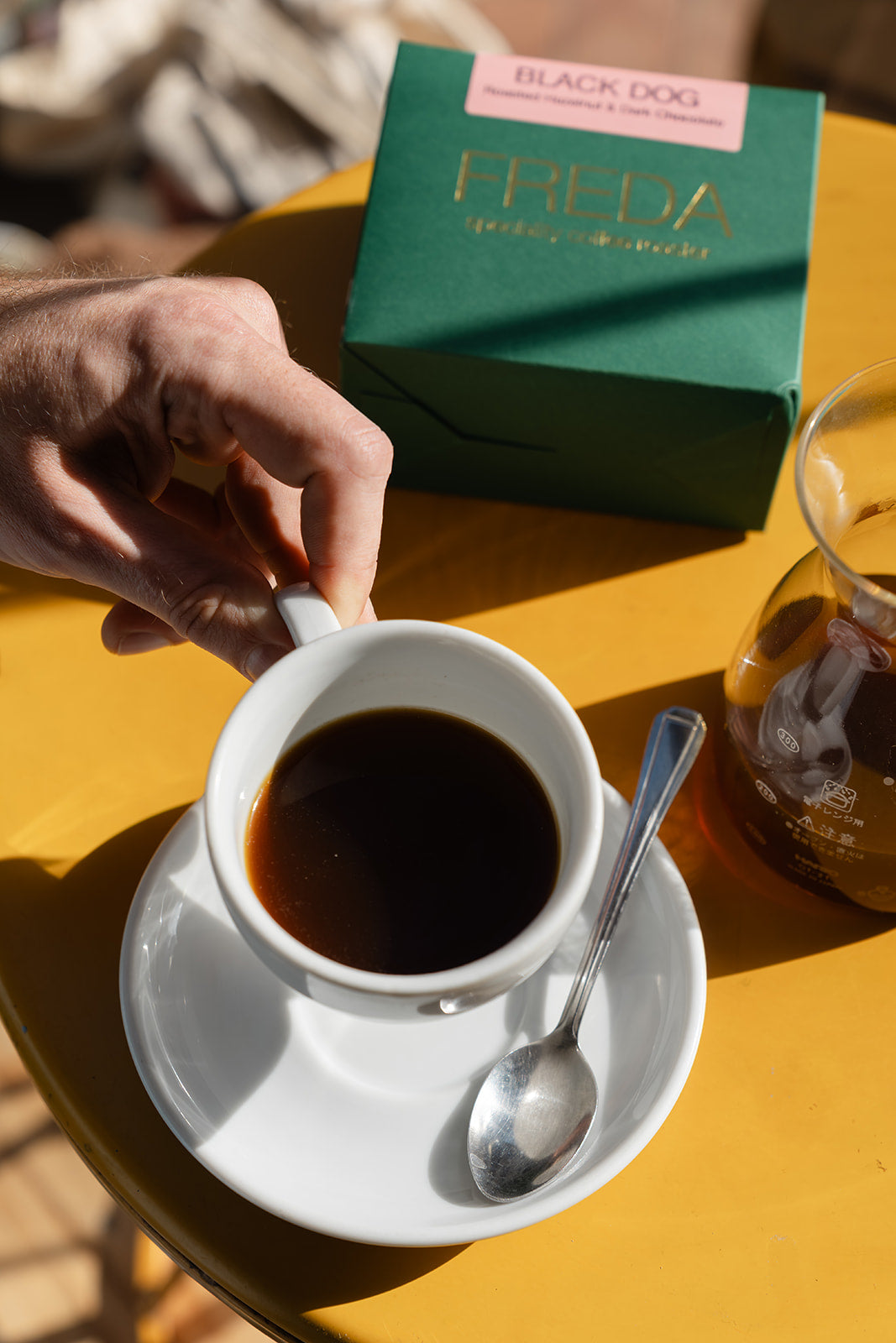 Coffee cup in Bond Street Coffee on a yellow table, with a Freda Speciality Coffee box in the background.