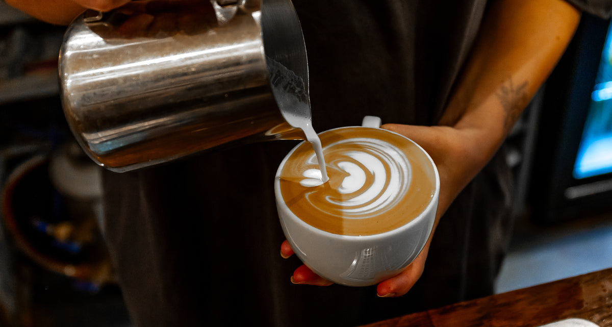 A close-up of a barista pouring steamed milk from a stainless steel pitcher into a white cup of coffee with latte art, set in a speciality cafe environment in Brighton
