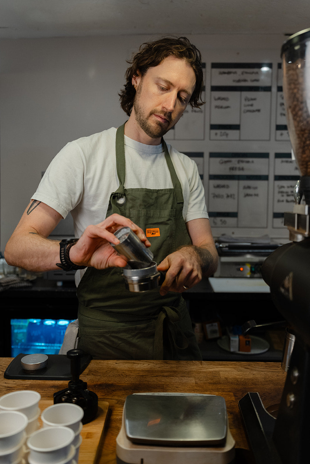Barista pouring ground coffee from a metal dosing cup into a portafilter fitted with a funnel.