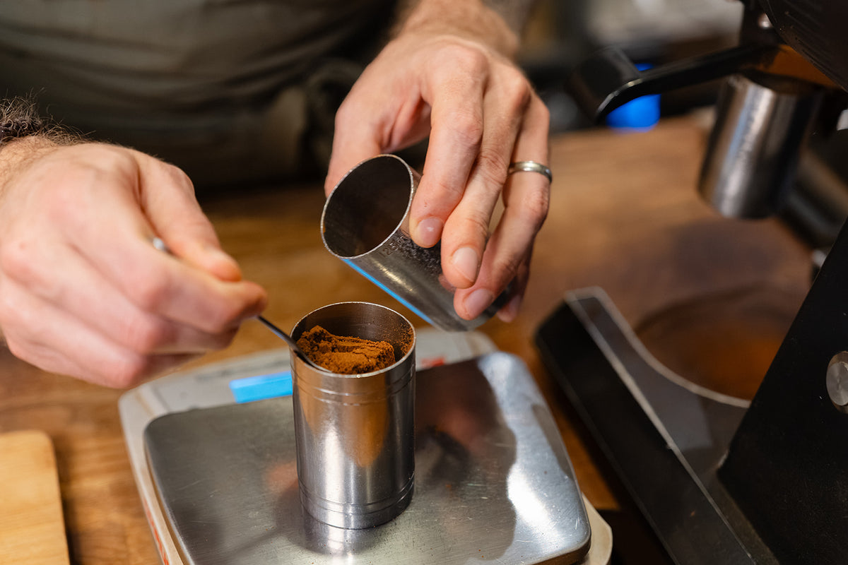 Barista adjusting the weight of ground coffee in a metal dosing cup on digital scales.